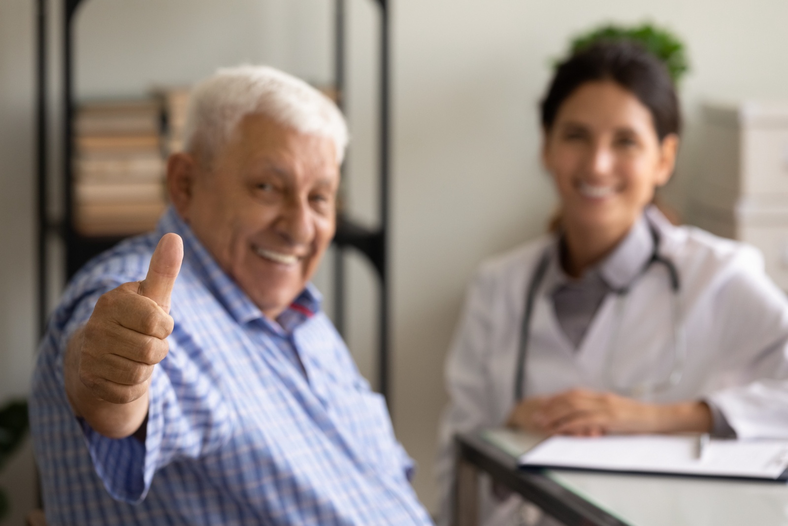 Close up focus on hand mature patient showing thumb up, female therapist and senior man sitting in office, happy old client satisfied by health insurance service, good healthcare concept
